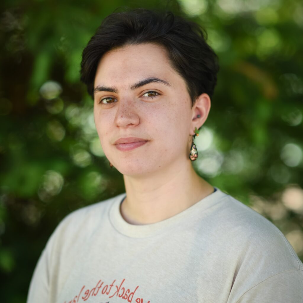 A photo of Lau outside. They are wearing a beige shirt with the text "give back to the land," part of which can be seen in the photo. Behind them are the blurred green leaves of a tree. 