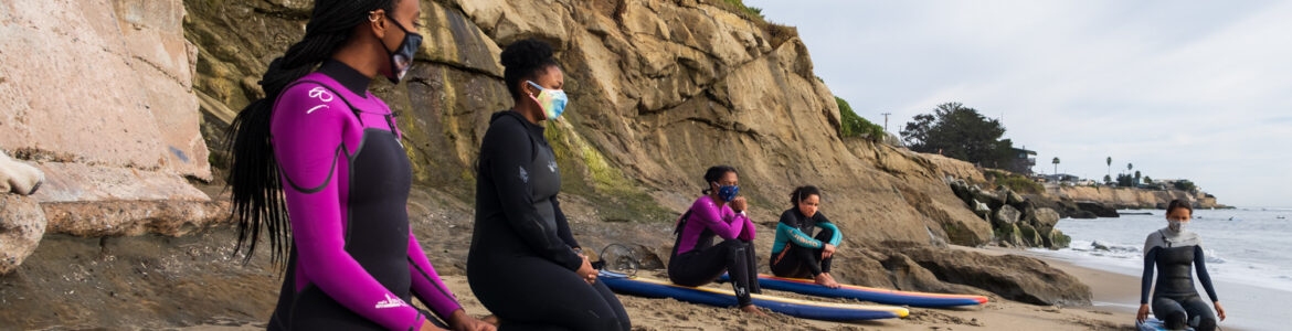 Four Black people on surf boards on the beach. They are wearing surfing attire and faced by another person who seems to be their instructor.