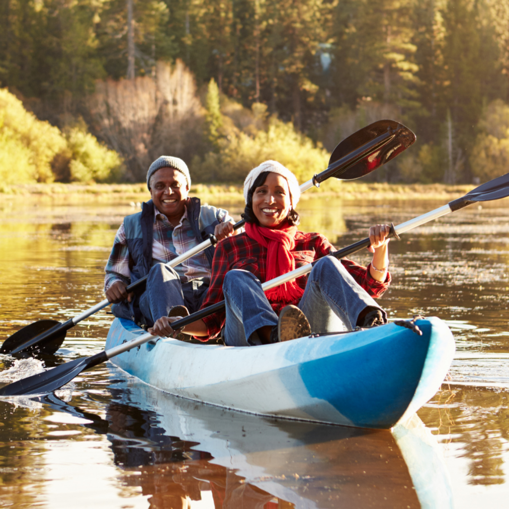 Two people in their forties or fifties are smiling as they pose for a photo inside a canoe. The water around them is brilliant with fall colors reflected from the trees in a distance. 