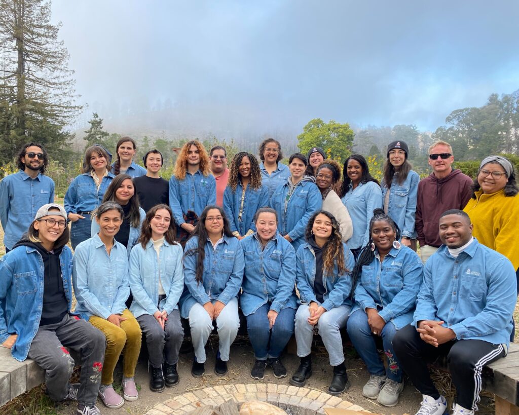 Justice Outside staff pose for a photo around a fire pit. There are trees and fog in the background. Most staff are wearing denim blue button up shirts with the organization's logo on them. 