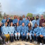 Justice Outside staff pose for a photo around a fire pit. There are trees and fog in the background. Most staff are wearing denim blue button up shirts with the organization's logo on them.