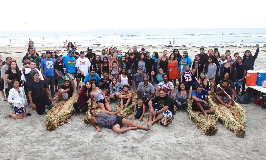 A large group of people of different ages are posing near the beach. They have with them four boats made of plant material. They are smiling at the camera.