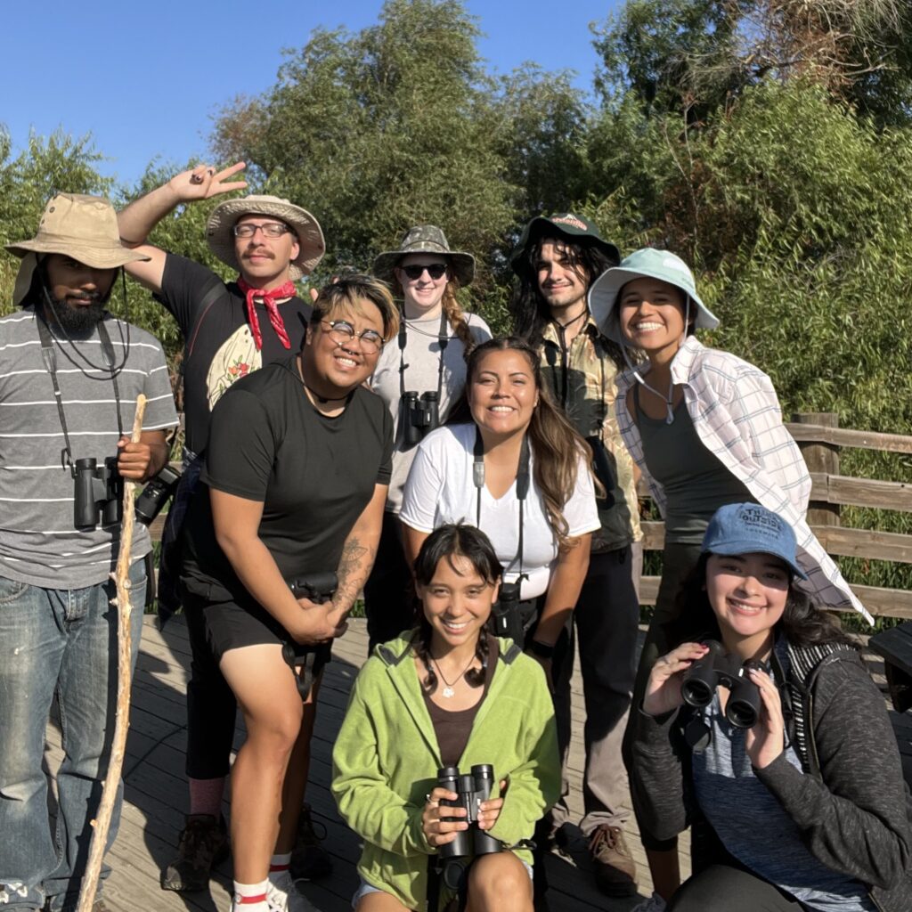 A group of mostly Young People of Color pose in front of lush trees outdoors. Some are holding binoculars. Many are wearing hats. 