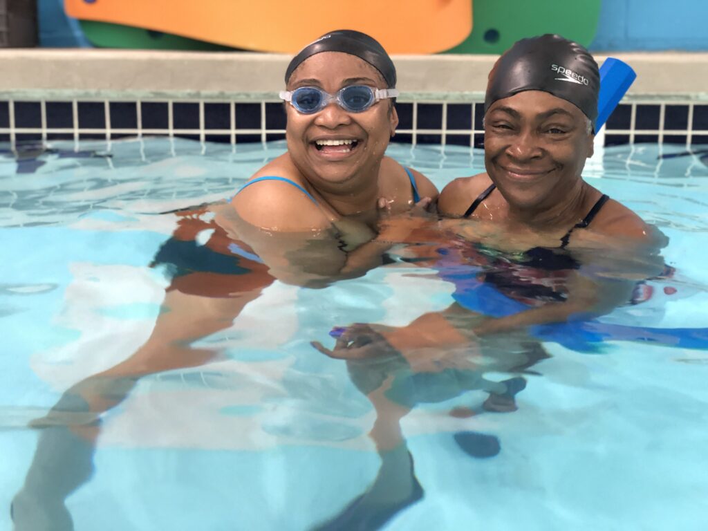 A mother and daughter are smiling in a swimming pool. The daughter is wearing goggles and they are both wearing hair caps. They have a blue pool noodle. 