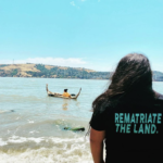 A person faces the water. The back of their t-shirt reads 'REMATRIATE THE LAND.'