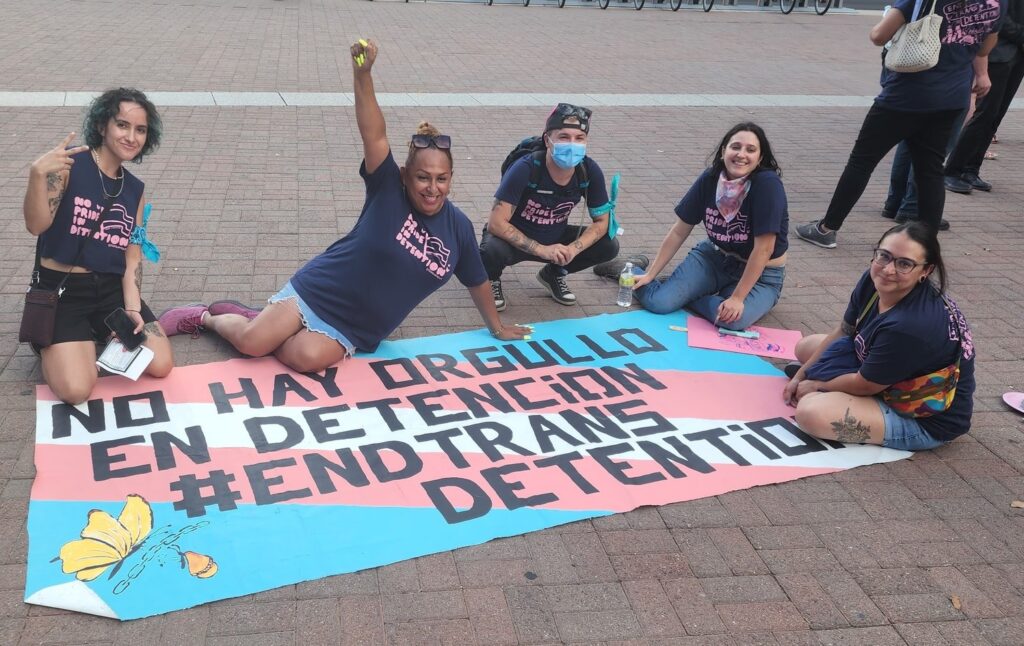 A group of five people are sitting on pavement with a sign next to them that reads "No Hay Orgullo En Detencion #EndTransDetention." The sign is made of the trans flag colors of white, pink, and blue. 