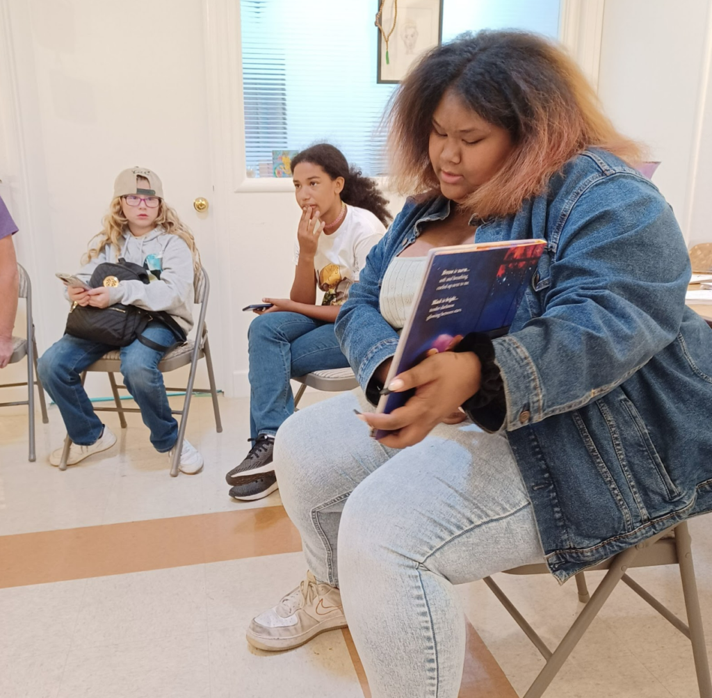 Three young people seated inside a classroom. One is holding a book and the other two holding cellphones and are in conversation. 