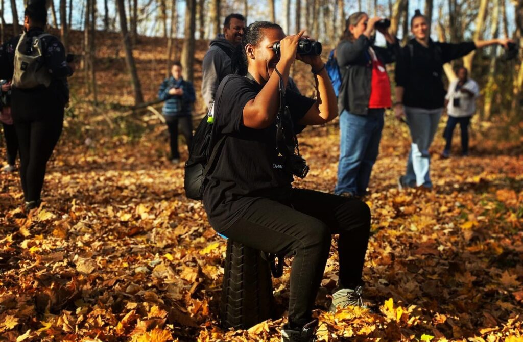 A group of people stand outside in the woods. Some are pointing in different directions. At the forefront a femme-presenting Black woman is sitting down and looking through binoculars. They are wearing all black. 