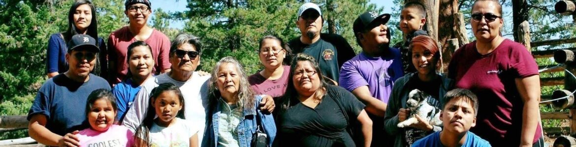 members of Diné Nihi Keyah Project family stand outside with trees surrounding them. They vary in age and are huddled near each other, embracing one another
