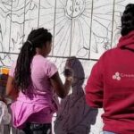 Five people, including at least two children with their backs to the camera as they paint a mural.