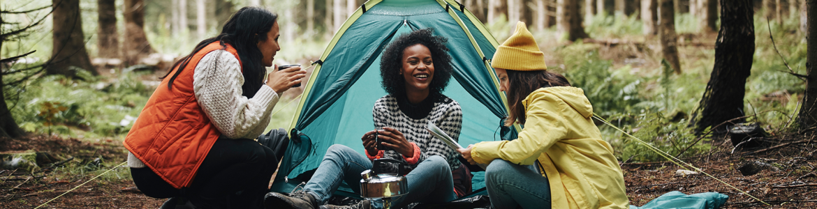 Three people sitting in the woods near a green tent. One is holding a map and another a mug of tea or coffee while the person in the middle smiles.