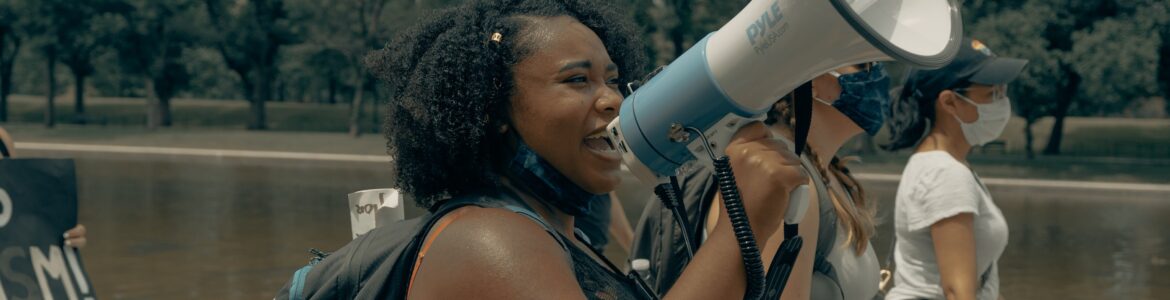 Image of a Black femme person holding a loud speaker and marching near a body of water with two other people.