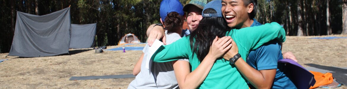 Four people hugging outdoors. There's a tent behind them.