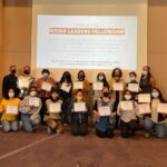 Group photo of facilitators with some of our Rising Leaders Fellows holding up their certificate of completion. Behind them is a screen projected that reads, "Thank You Rising Leaders Fellowship" and their names listed below.