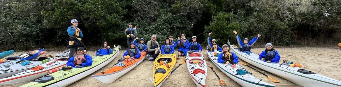 A group of young people, most of them people of color, on kayaks near water and smiling at the camera.