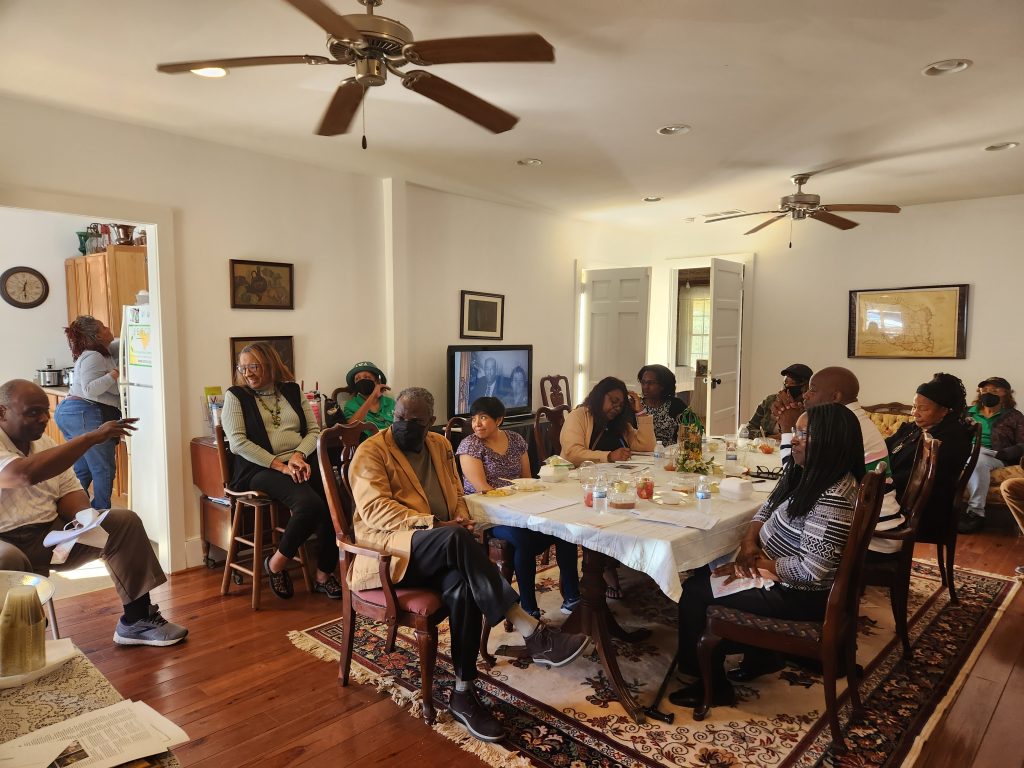 A group of more than a dozen people sit around a dining table together. Many are wearing masks. A masculine Black leader is speaking with the people in the room and holding a piece of paper. Folks are smiling. 