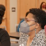Three femme presenting folks sitting at a conference table wearing masks.