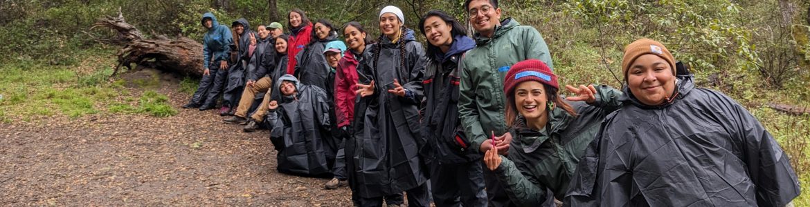A group of young people standing outside in rain jackets. They are smiling and looking at the camera, surrounded by greenery.