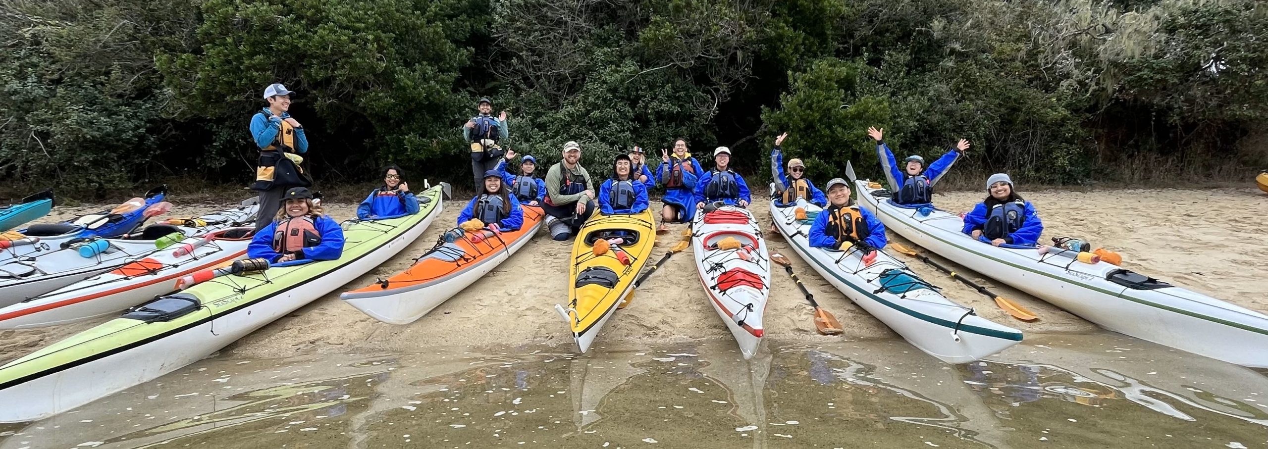 A group of young people of color on canoes near a body of water. They are wearing life jackets and smiling at the camera. 
