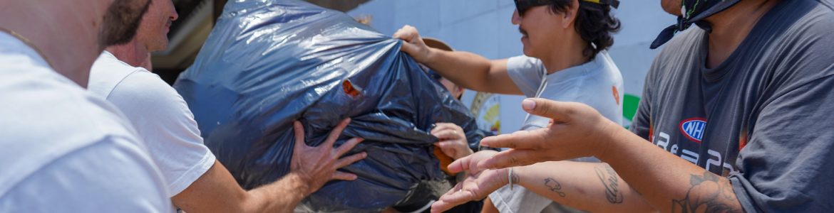 Four people are standing outside, passing down a plastic bag filled with goods. They are all wearing hats and t-shirts.