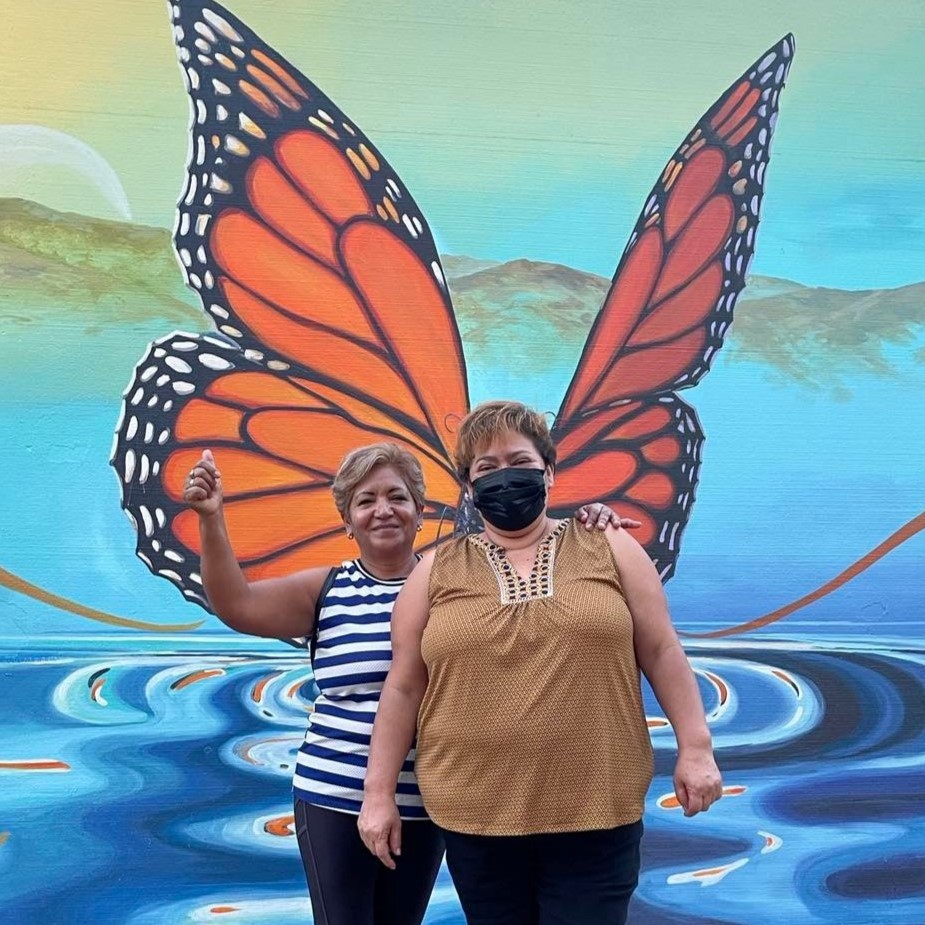 Two people standing in in front of a monarch butterfly mural. The wings of the butterfly are behind them as if they have wings. The person in the front is wearing a taupe shirt and black pants and a black fast mask. The person in the back is wearing a stripped blue and white shirt with black pants and has their fist up in a celebratory/active gesture. They both have short hair.