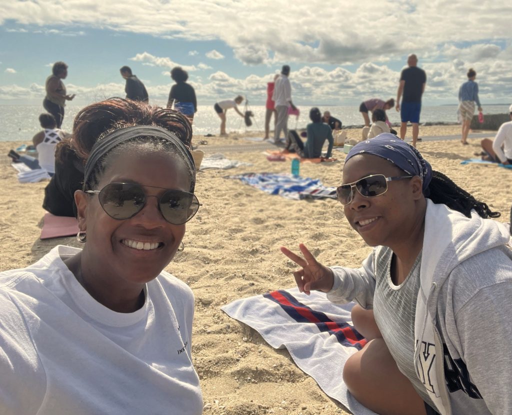 Kim and her friend Rebecca on the beach. They are both wearing sunglasses and light colored shirts and looking at the camera. Behind them, there are other people enjoying the beach and white fluffy clouds in the sky.