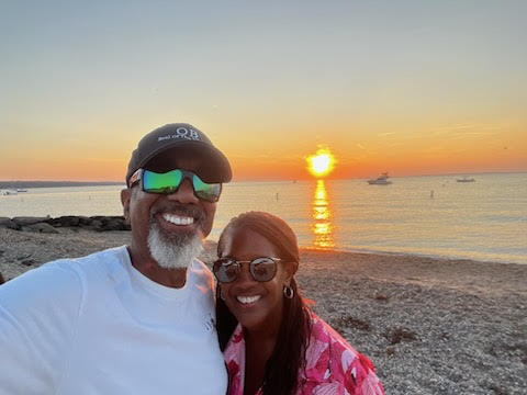 Kim and her husband Mike on the beach during sunset. They are both smiling and wearing sunglasses. 