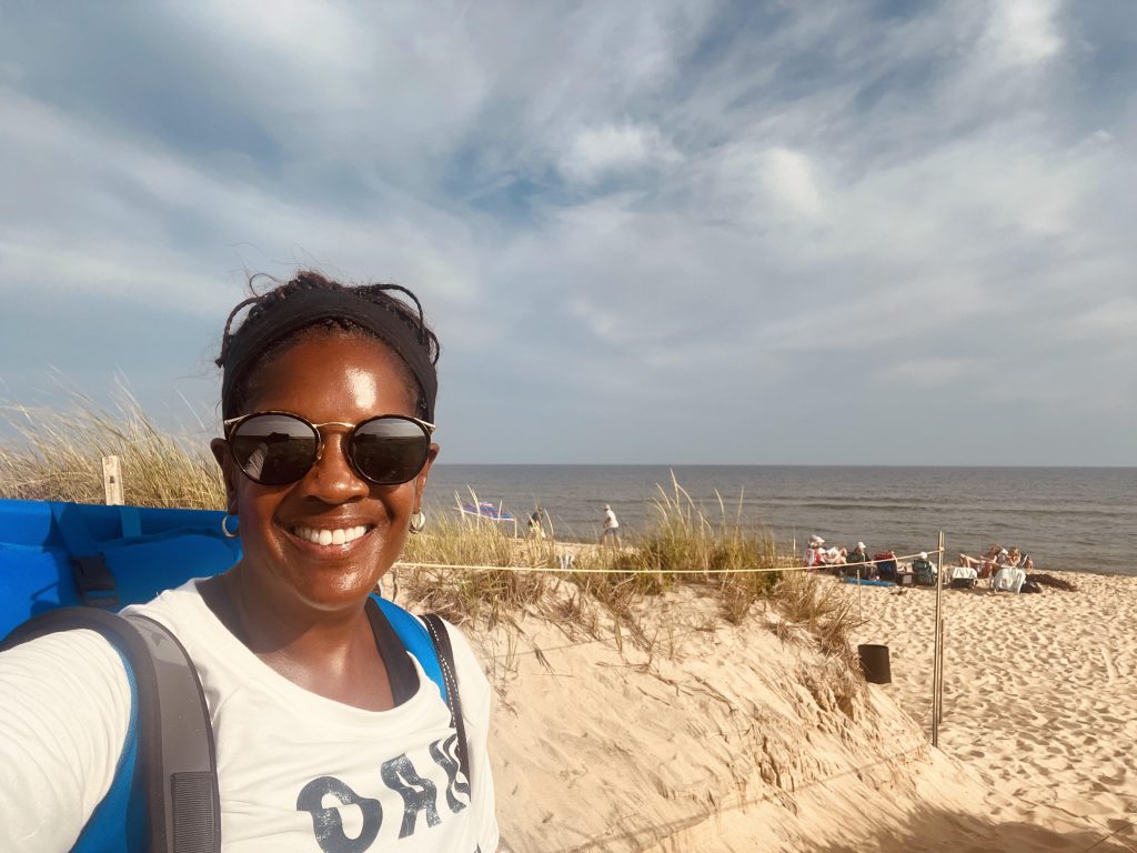 Kim on the beach, wearing a white shirt and sunglasses and a backpack. Looking at the camera. 