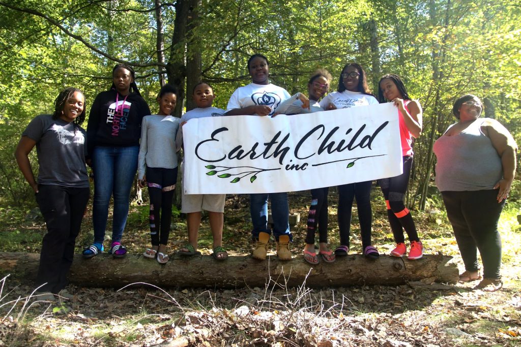A group of nine children and adults stand outside with a sign that reads "Earth Child inc." They are surrounded by trees.