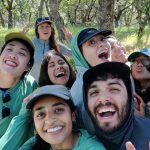 A selfie of a group of young people, most of them people of color, looking at the camera, smiling, laughing and cheering while they stand outside among trees.
