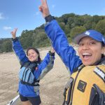 Two participants in Outdoor Educators Institute stand outside on a beach. They are wearing blue and life vests and they have their hands up in the air in celebration.