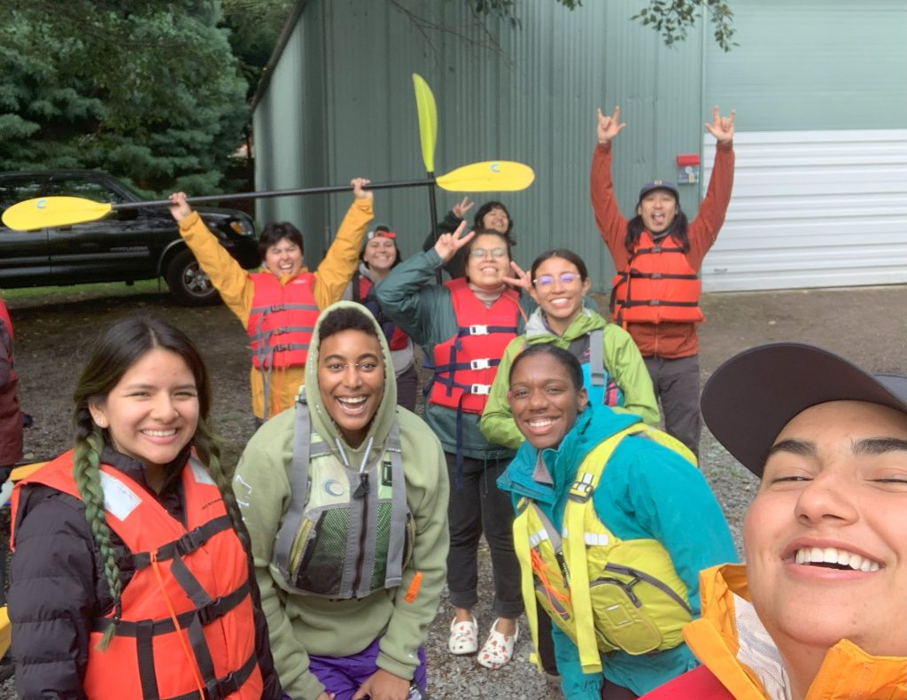 A group of 10 Outdoor Educators Institute: Seattle participants stand outside. They are smiling and looking joyful. Many are wearing lifejackets and some are holding canoe paddles. 